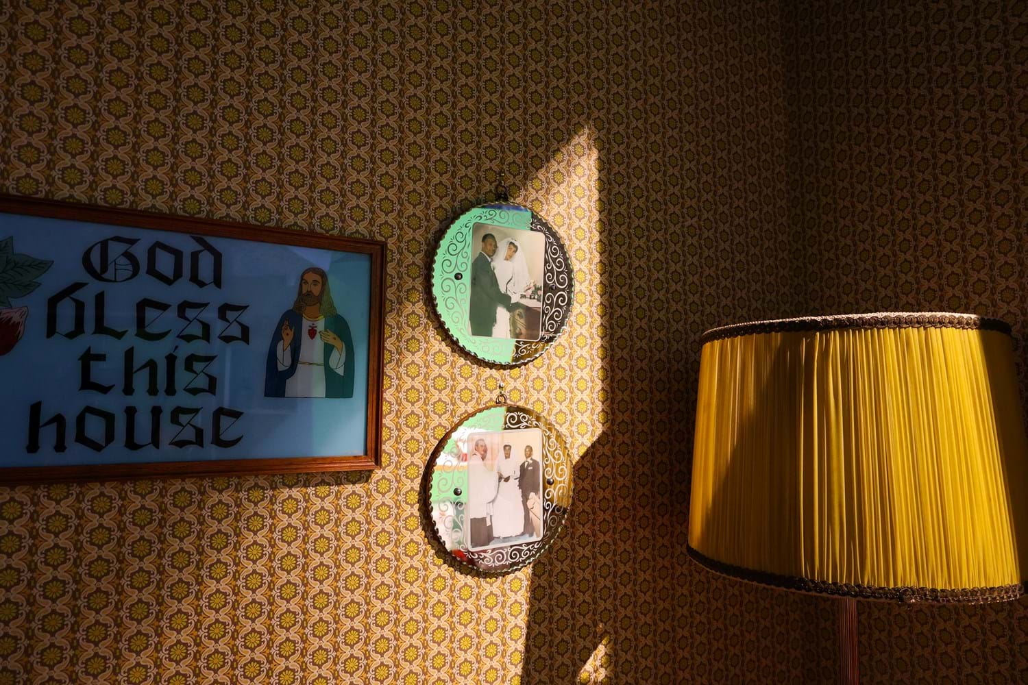 Two photos hanging on a wall dressed with seventies patterned wallpaper, a yellow lamp shade is on the right and a framed sign with the words "God bless this house" is on the left.