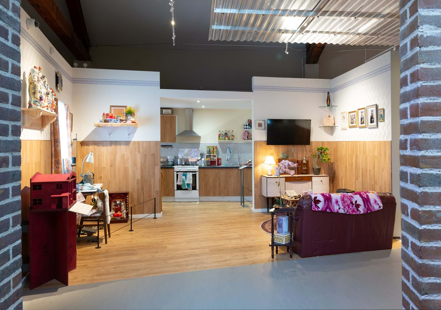 A wide shot of A Terraced House in 2024, the one of the rooms in Rooms Through Time at Museum of the Home. It's a living room and kitchen space for a British-Vietnamese family. The image includes different periodic & cultural items including different spices and ingredients in the kitchen, a sewing machine, a TV with karaoke on, etc.