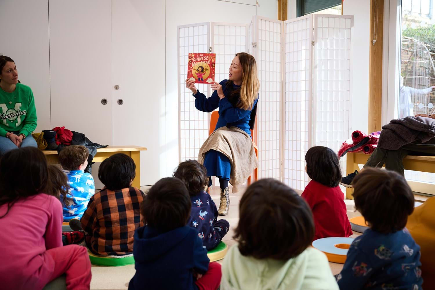 A woman is holding a children's book in a storytelling session, surrounded by children.