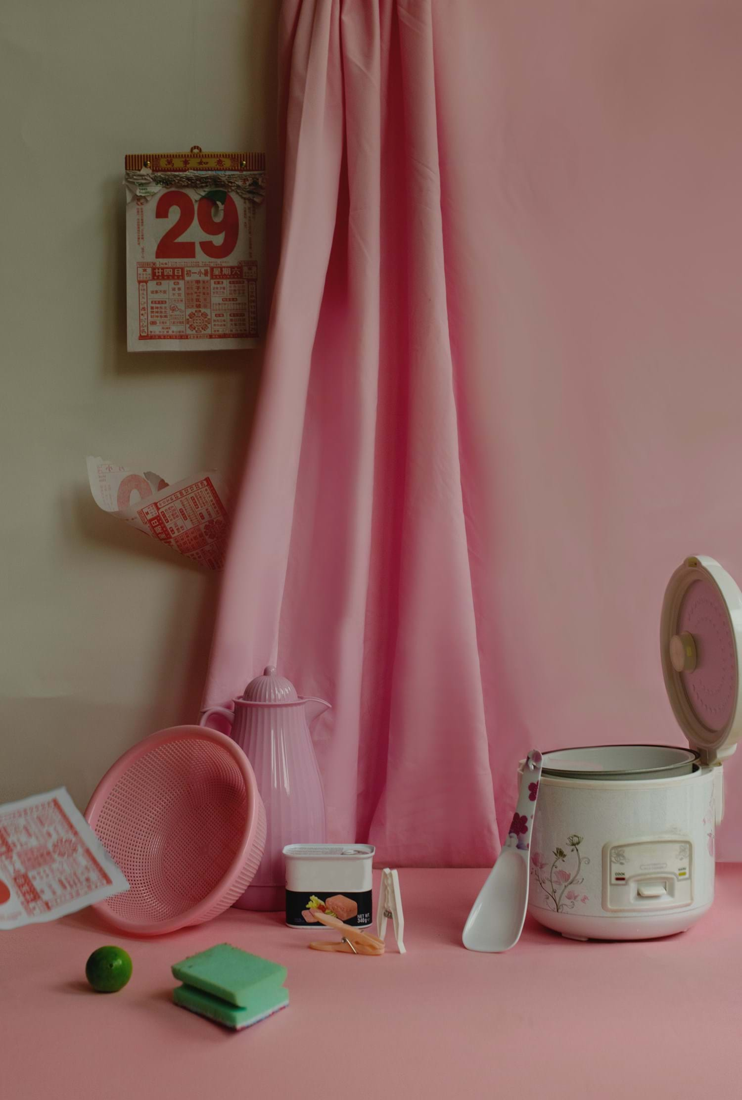 A pink back drop and pink curtain hanging with kitchen elements including a rice cooker, sponge and tear away calendar