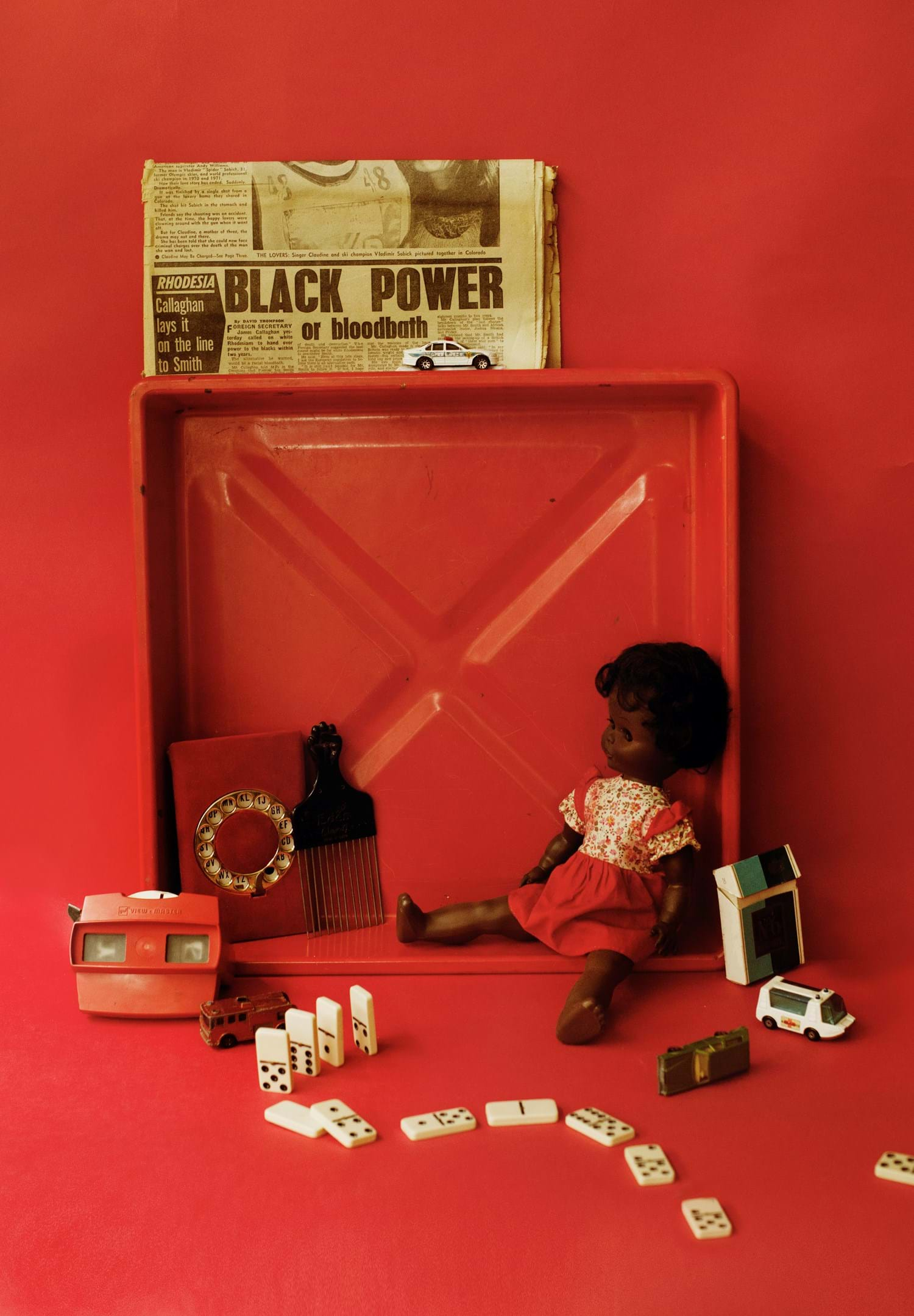 A red backdrop and vignette with a children's doll, hair comb, dominoes and newspaper in the forefront