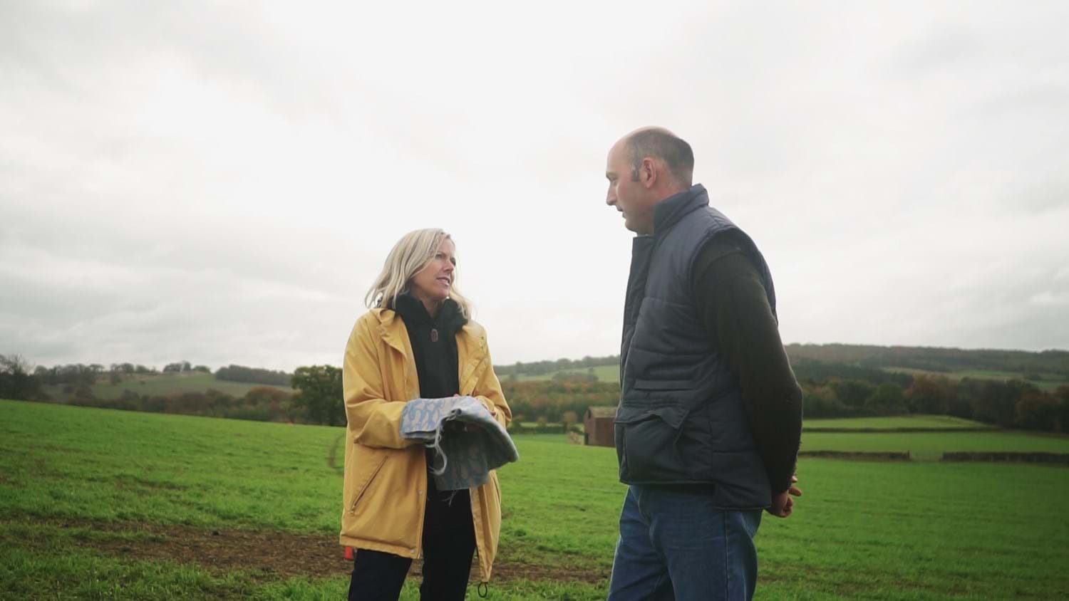 Ella Doran (left) is having a conversation with Farmer Platt (right) in a green open field. Doran is holding fabric in her hands.