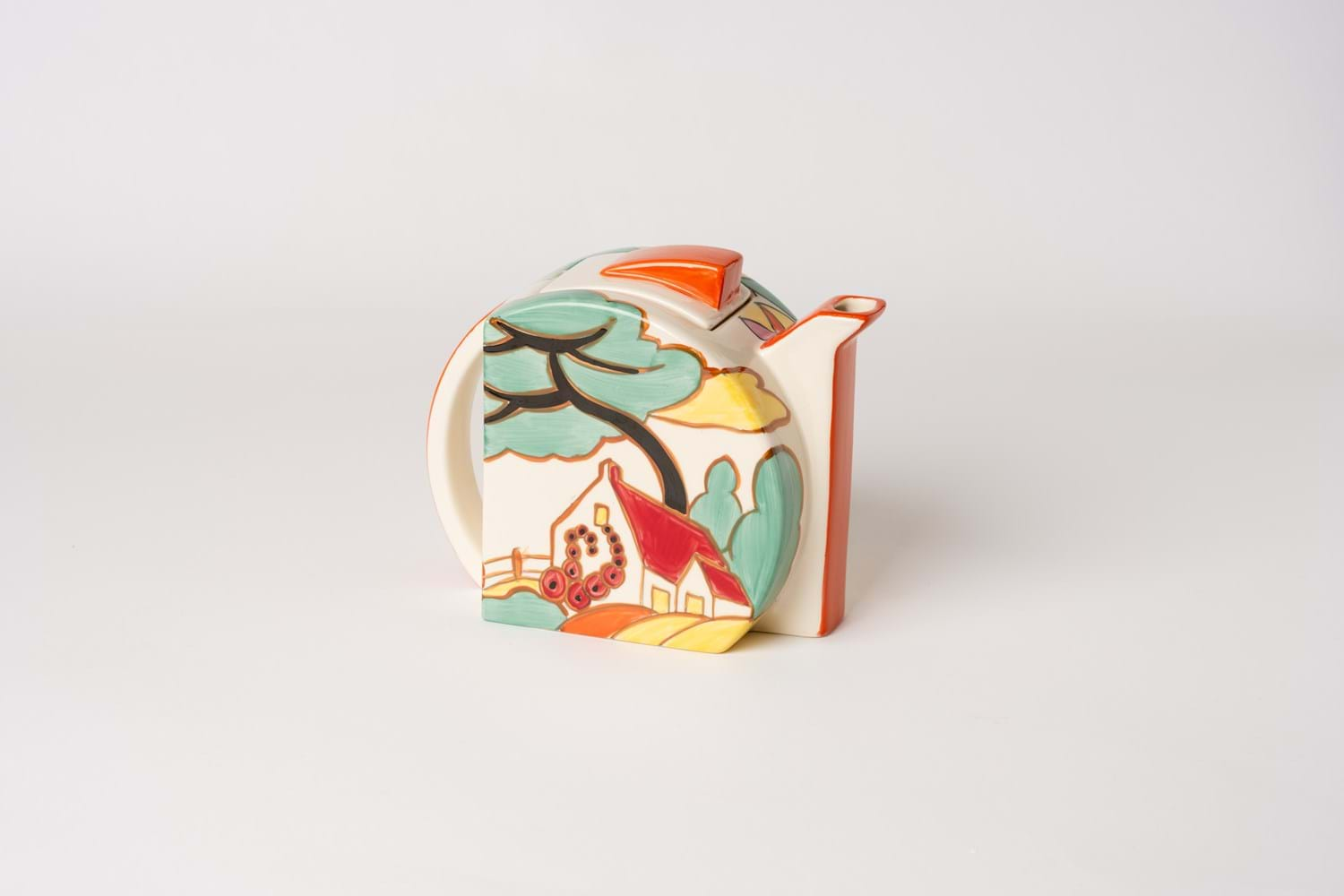 A small geometric teapot with colourful painted decorations depicting a house and tree