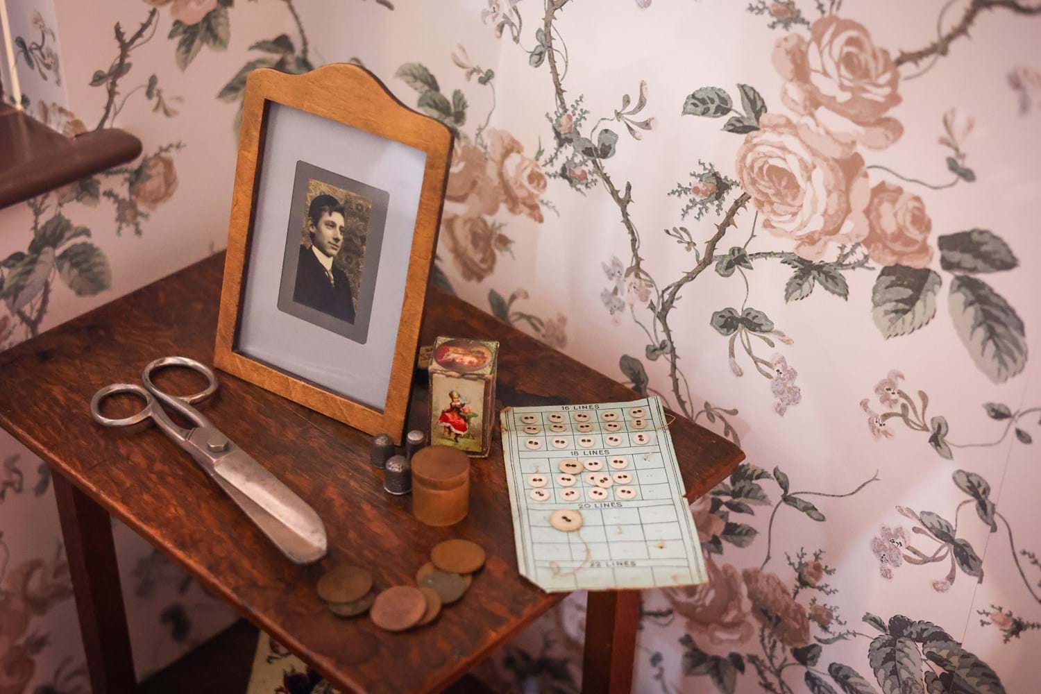 A wooden bookshelf against a wall with pink rose patterned wallpaper. Atop the shelf is a framed photo of a person in the 1910s, buttons, and sewing scissors