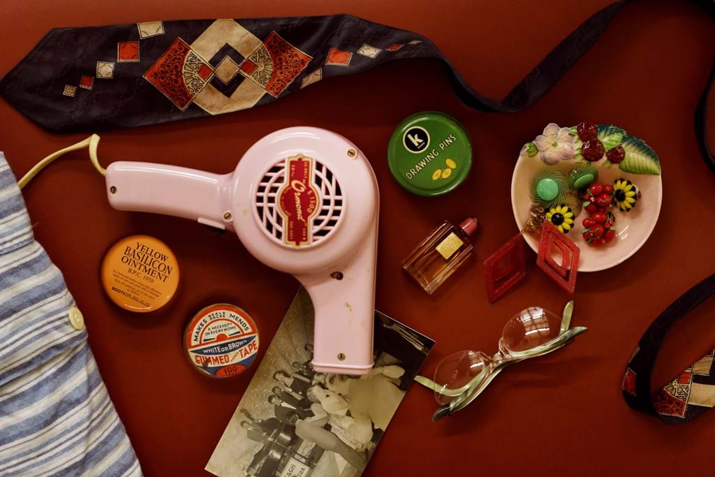 A dark red backdrop with objects from the fifties laid out flat, including a vintage pink hairdryer, blue patterned tie, cat-eye glasses frames and earrings in a pink dish