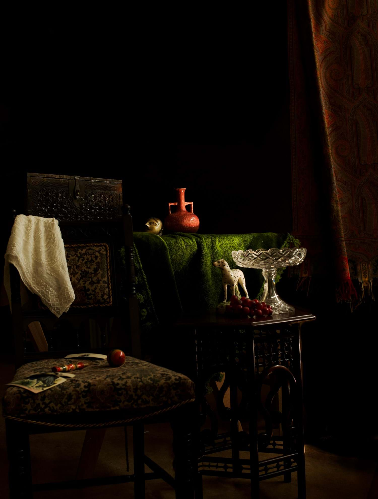 A corner of a room lit dramatically. Curtain on both sides and various furniture and home items from 1870s, such as chairs, a trinket, vase, fabrics, and more, stacked and gathered together.