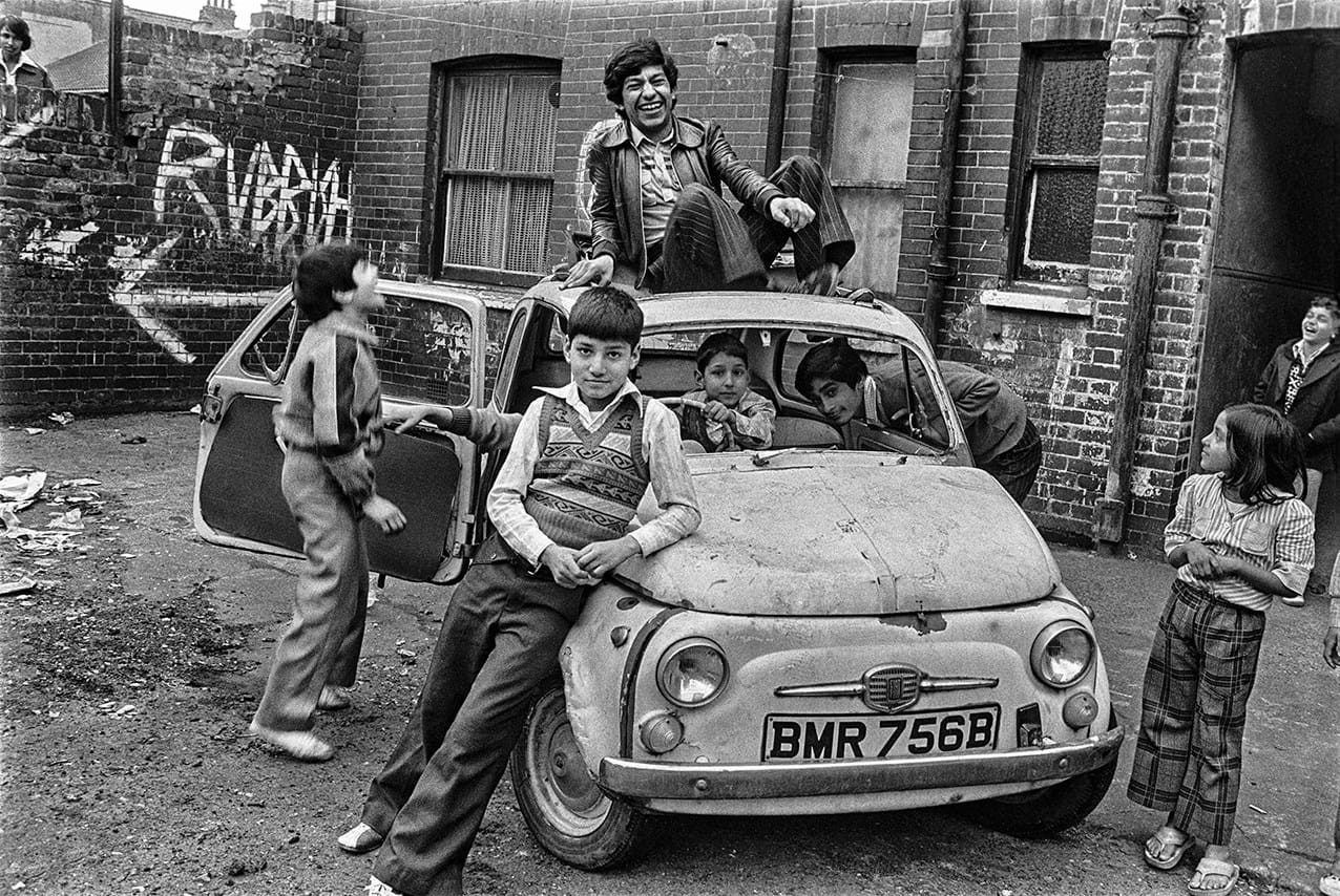 Black and white photograph of several young people smiling while leaning against, inside, and on top of a car 