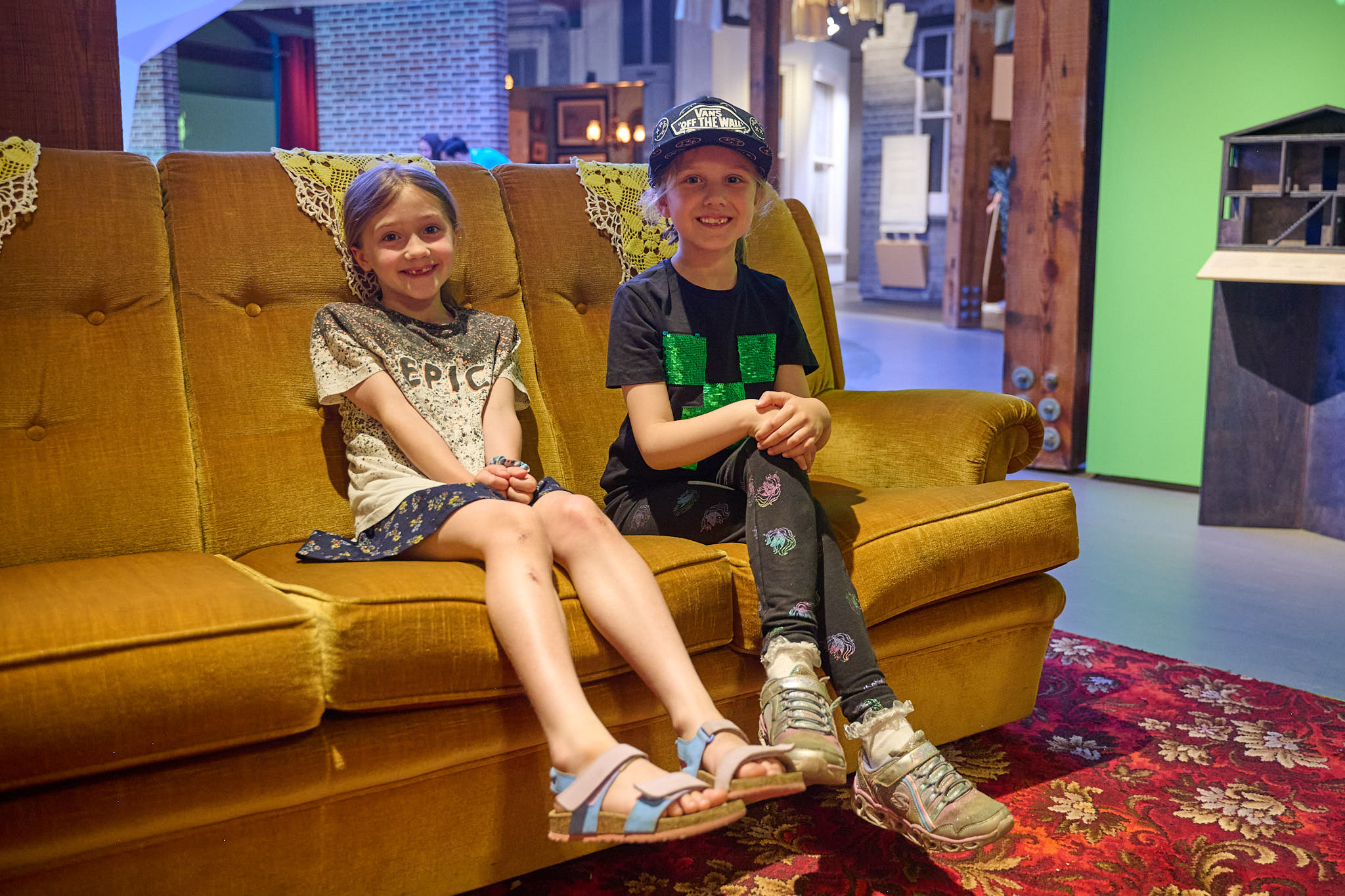 Two children are sitting side by side on a mustard yellow sofa draped with crocheted doillies on red floral patterned carpet. They are both smiling and looking directly into the camera.