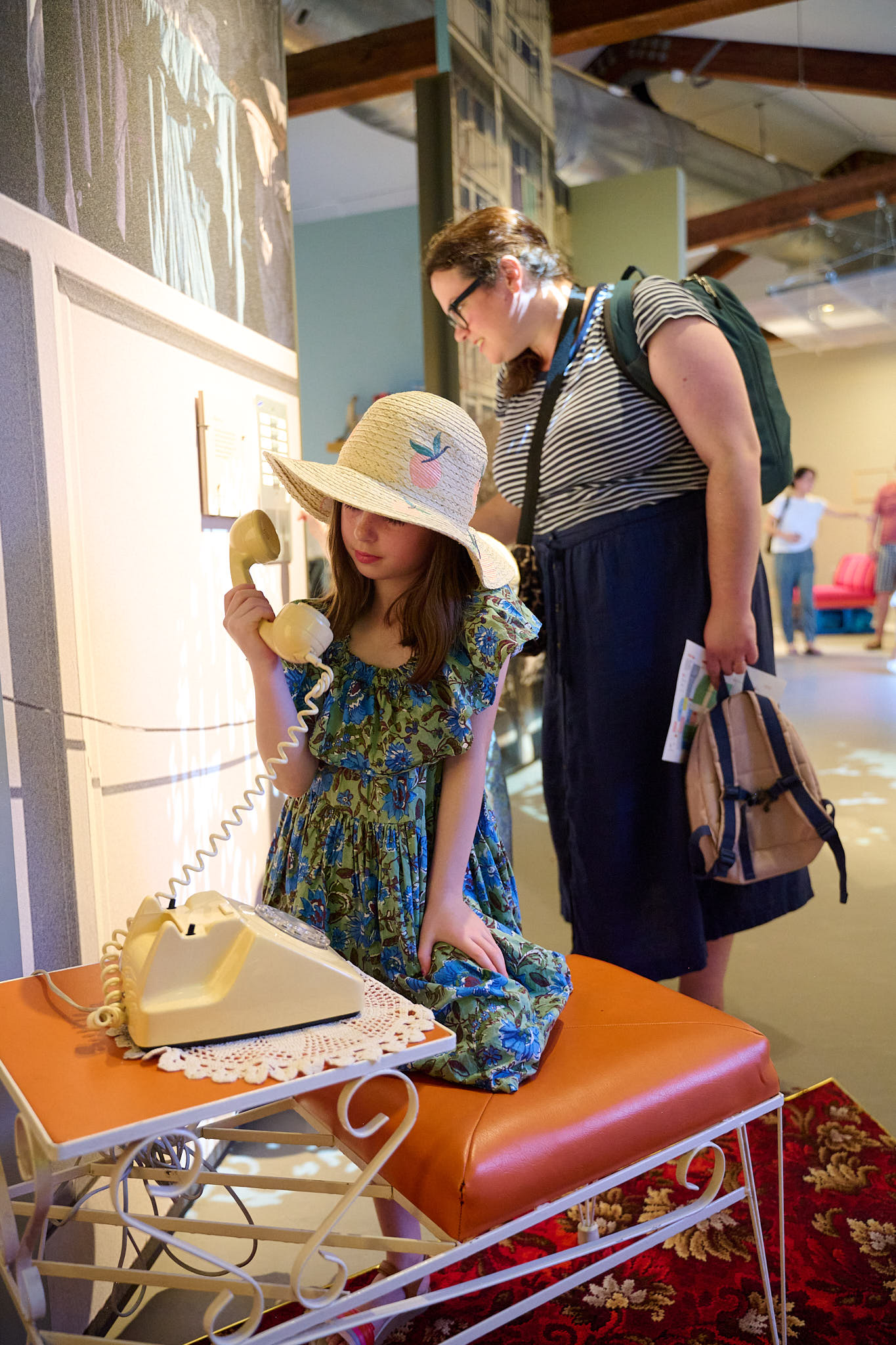 Two people interacting with the installation in Rooms Through Time. A child kneels on an orange padded chair holding the handset of a beige rotary telephone near her ear. Behind her, an adult leans forward to interact with a doorbell installation on the wall.