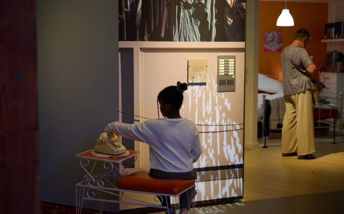 Two people interacting with the installation at the Museum. A child sits on an orange padded chair while holding the handset of the rotary telephone. In the back is an adult stands in a room with a bed in an orange wall, looking at something outside of the frame.