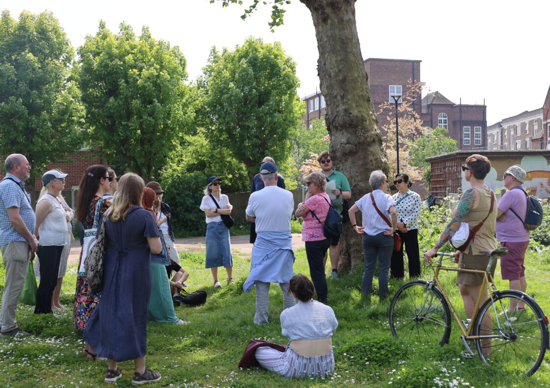 A group of people stand facing towards a tour guide in a park with grass and trees