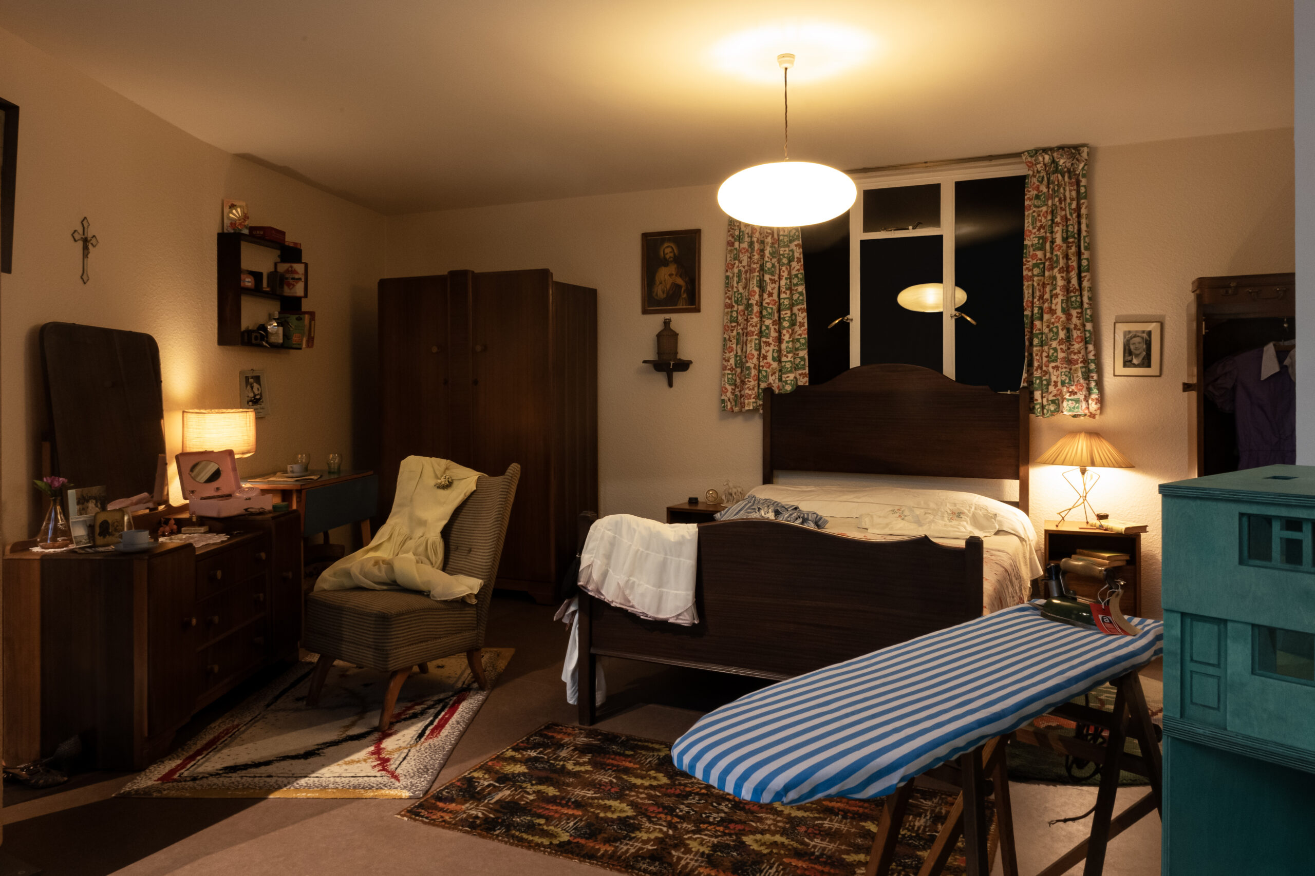 An installation view of A Room Upstairs in 1956 at the Museum. There are clothes scattered on the bed in the centre. A vanity area on the left is various items. On the right foreground is an iron board. There are a cross and religious painting on the wall. In the background, there is a closet with an NHS nurse uniform from the 1950s.