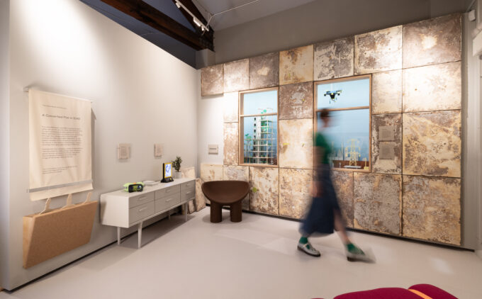 A blurred figure walks across a room in front of weathered looking panels and windows showing a model of a city.