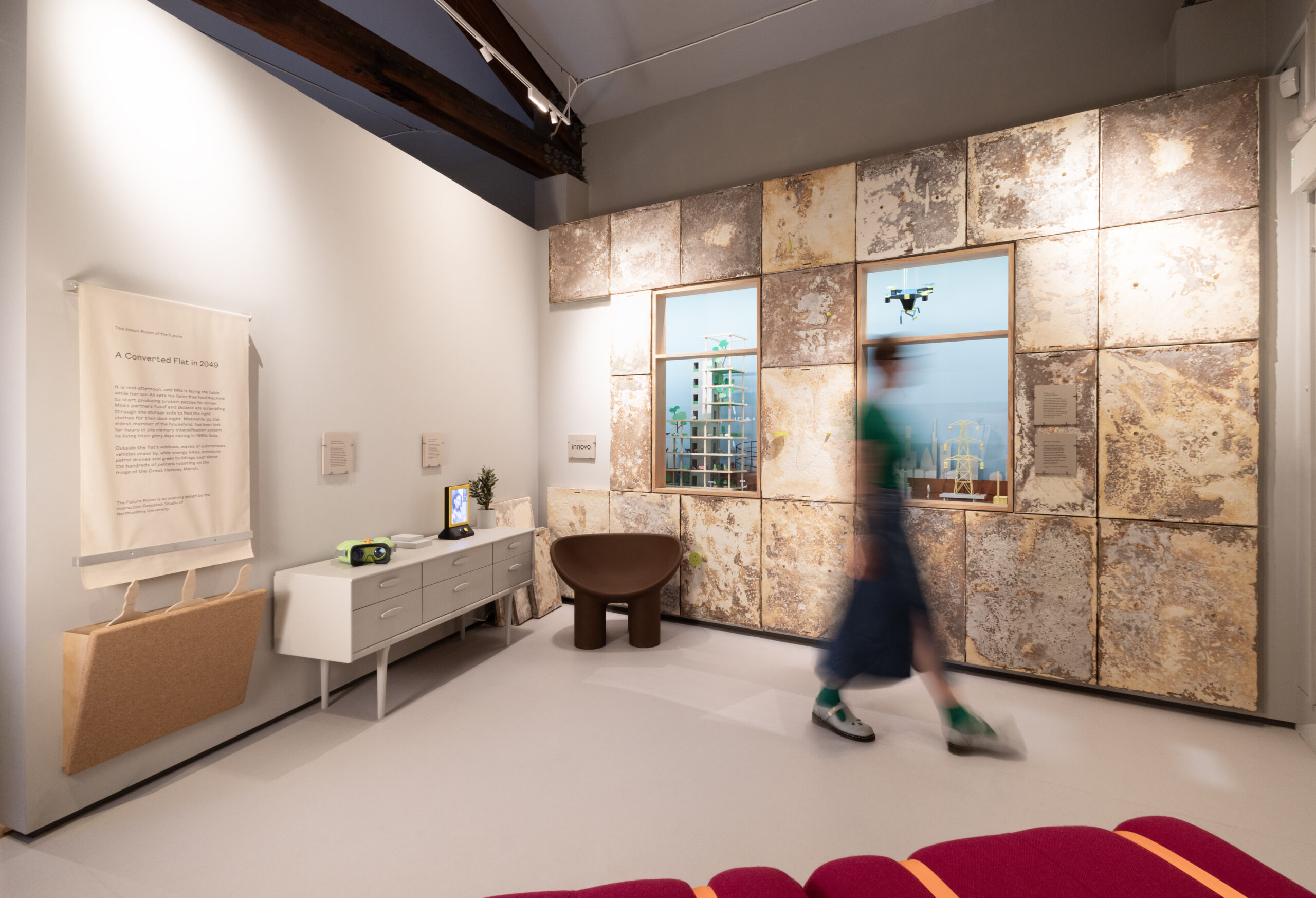 A blurred figure walks across a room in front of weathered looking panels and windows showing a model of a city.