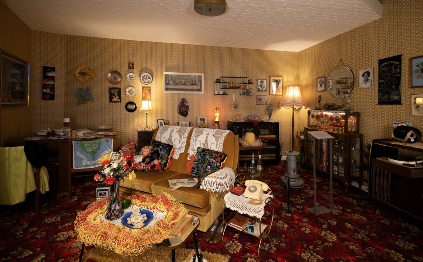 A Caribbean British Front Room installation. The floor in red floral patterned carpet with mustard yellow wallpaper. In the centre is a greenish gold two seater sofa is draped with white crocheted doillies and patterend cushions and dolls. There are various framed photos and decors on the wall, cabinets, and coffee table.