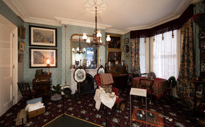 An installation view of A Townhouse in 1878 at the Museum. A deep red carpet and mint green patterned wall paper room holds framed artworks on the wall, fireplace and a mirror on the mantel, table with wooden chairs, writing desk, and piano. On the floor are children's toys and dolls as well as an ayah's trunk full of textiles.