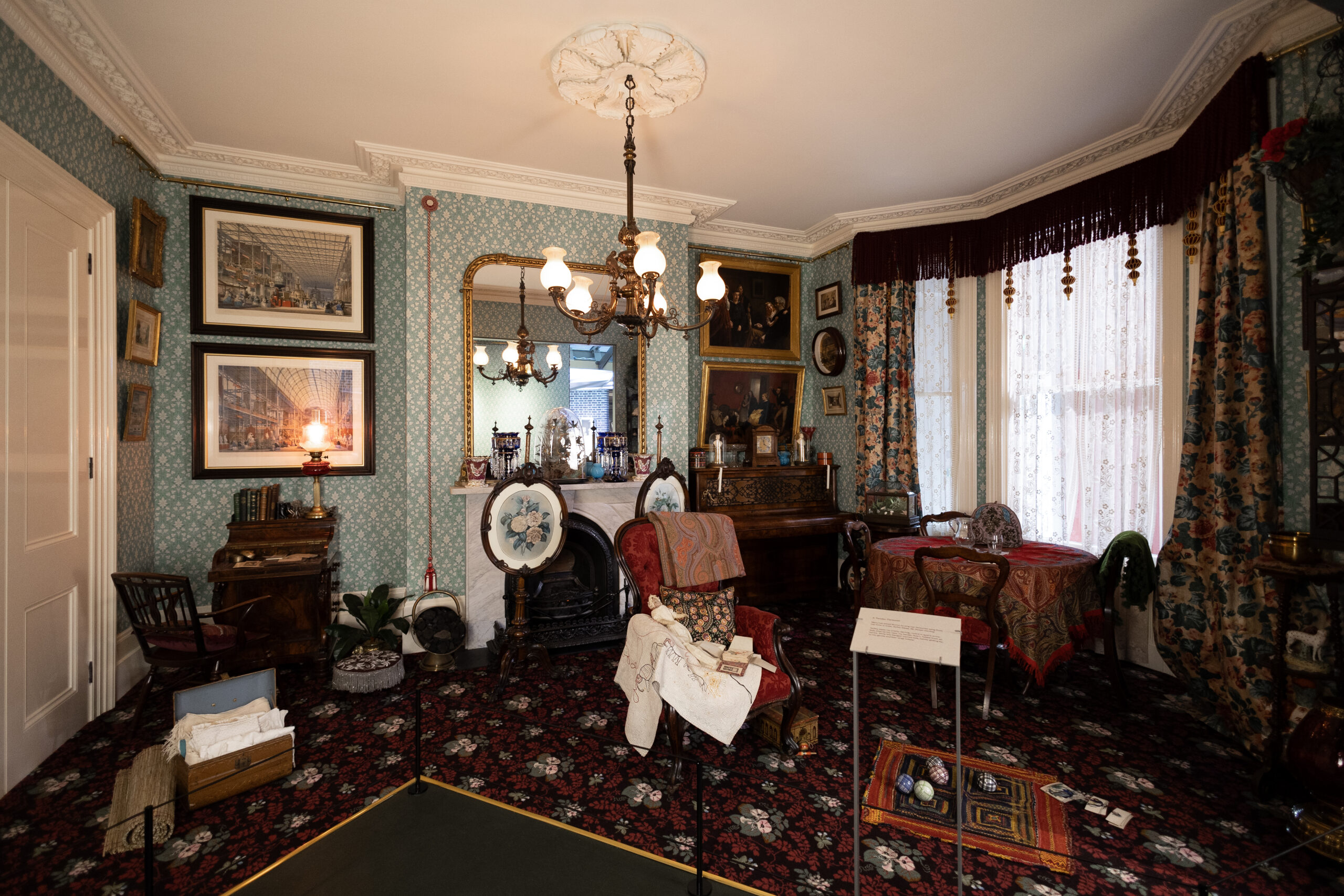An installation view of A Townhouse in 1878 at the Museum. A deep red carpet and mint green patterned wall paper room holds framed artworks on the wall, fireplace and a mirror on the mantel, table with wooden chairs, writing desk, and piano. On the floor are children's toys and dolls as well as an ayah's trunk full of textiles.