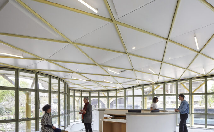 A bright, airy reception space with a gently curved white desk at centre right, behind which a staff member speaks to a visitor. To the left, a large semi-circular bank of green-framed windows looks out onto leafy gardens. The person on a bench on the left is chatting with a person standing right to the bench.