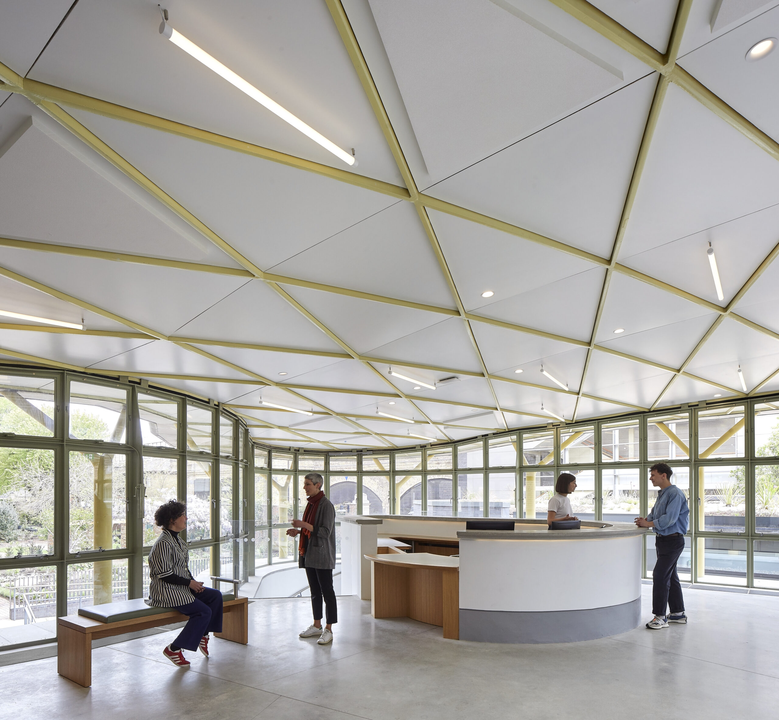 A bright, airy reception space with a gently curved white desk at centre right, behind which a staff member speaks to a visitor. To the left, a large semi-circular bank of green-framed windows looks out onto leafy gardens. The person on a bench on the left is chatting with a person standing right to the bench.