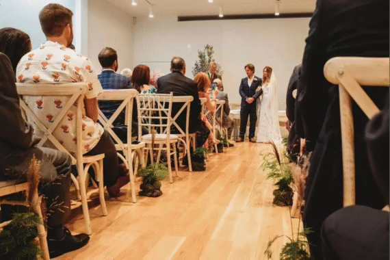 Indoor wedding ceremony with guests seated on wooden chairs watching the bride and groom exchange vows in a modern white-walled venue.