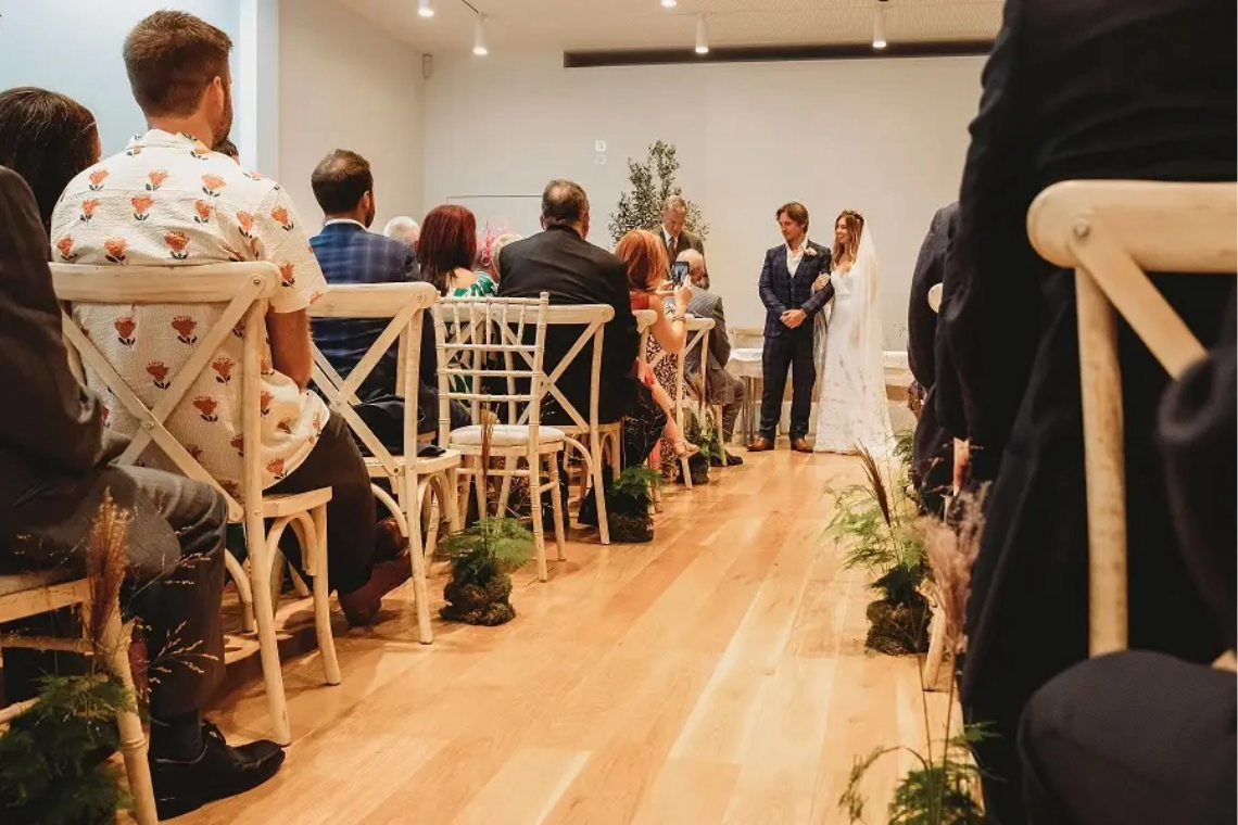 Indoor wedding ceremony with guests seated on wooden chairs watching the bride and groom exchange vows in a modern white-walled venue.