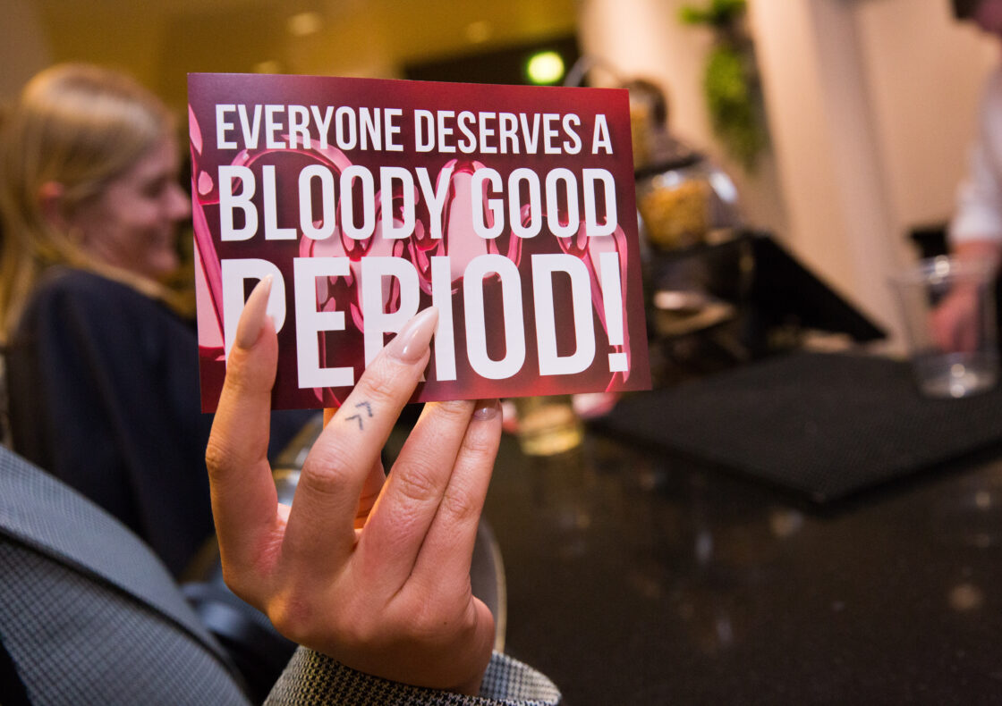 Close-up of a hand holding a sign reading ‘Everyone deserves a bloody good period!’ against a background of people socialising at an indoor event.