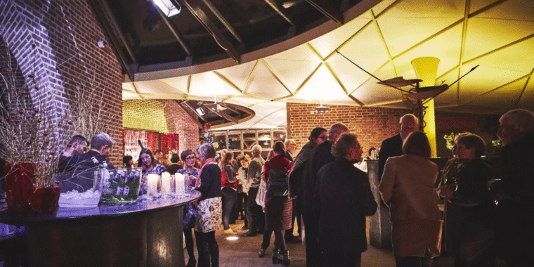 Crowded evening event in a brick-walled venue with dramatic lighting, featuring guests socializing around a curved bar area.