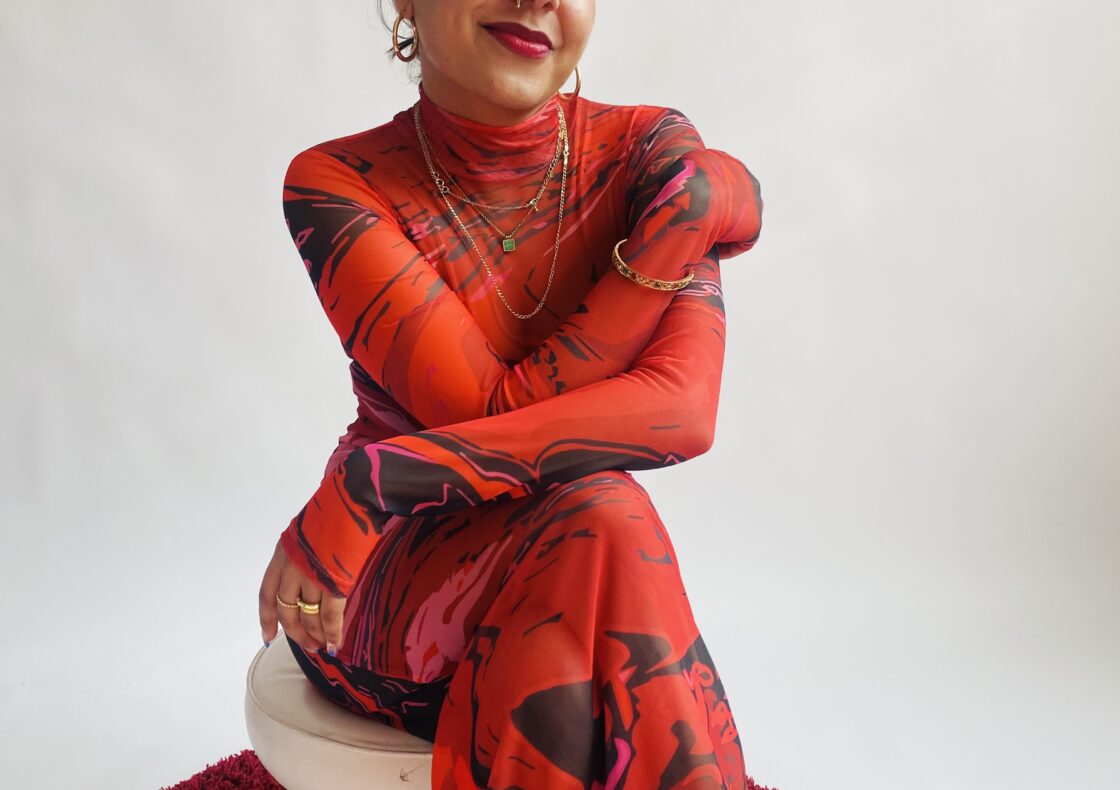 Person seated on a small white stool against a plain backdrop, smiling. They wear a sheer red patterned outfit with layered gold jewellery, hoop earrings, and black heels, seated on a red circular rug