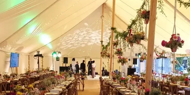 Wedding reception in a white tent decorated with hanging flowers and greenery, featuring long tables set for dinner with colorful lighting effects.