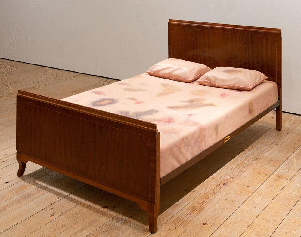 A wooden-framed double bed with peach-toned bedding and pillows, printed with faint, abstract patterns resembling bruises and body marks. Displayed in a minimal gallery space with wooden floors.