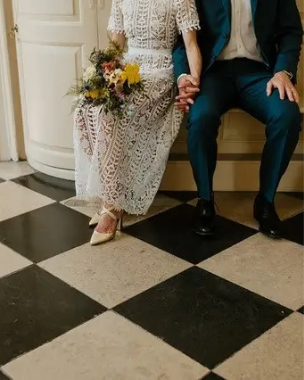 Two people hold hands on a bench in wedding attire.