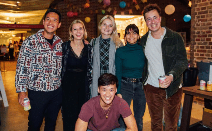 Group of six smiling young adults posing together indoors, holding drinks. Behind them in a distance is a brick wall decorated with colourful hanging lanterns.