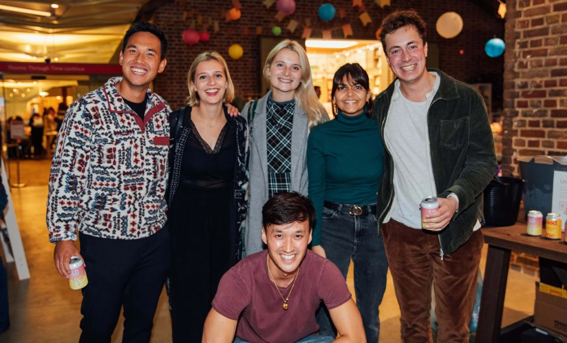 Group of six smiling young adults posing together indoors, holding drinks. Behind them in a distance is a brick wall decorated with colourful hanging lanterns.