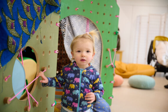 A small child looks at the camera as they play in a green wooden modular house