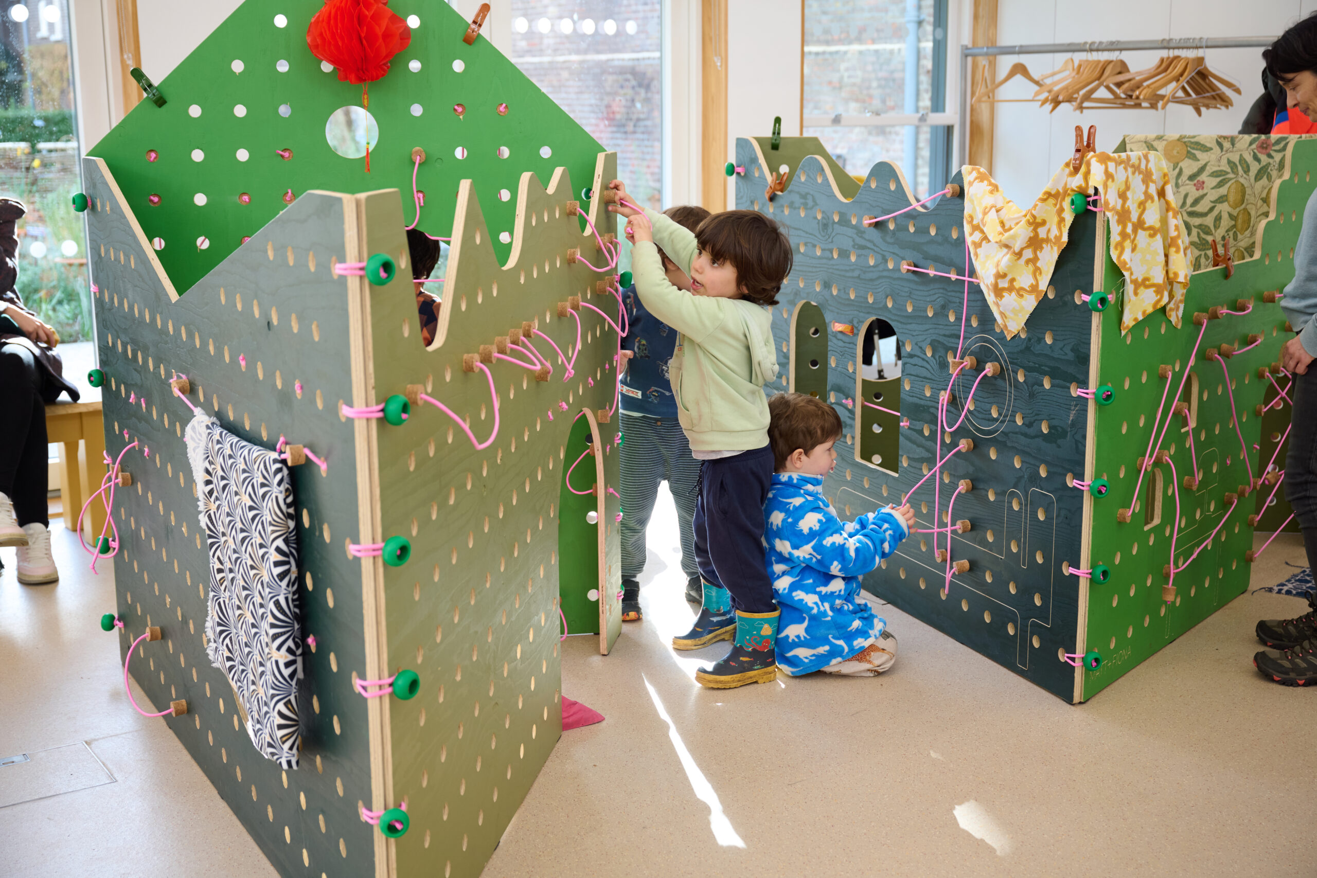 Three young children engaging with a large, green, wooden peg-board playhouse structure. Each interacting with different parts of the installation including pink elastic and bright green knob.