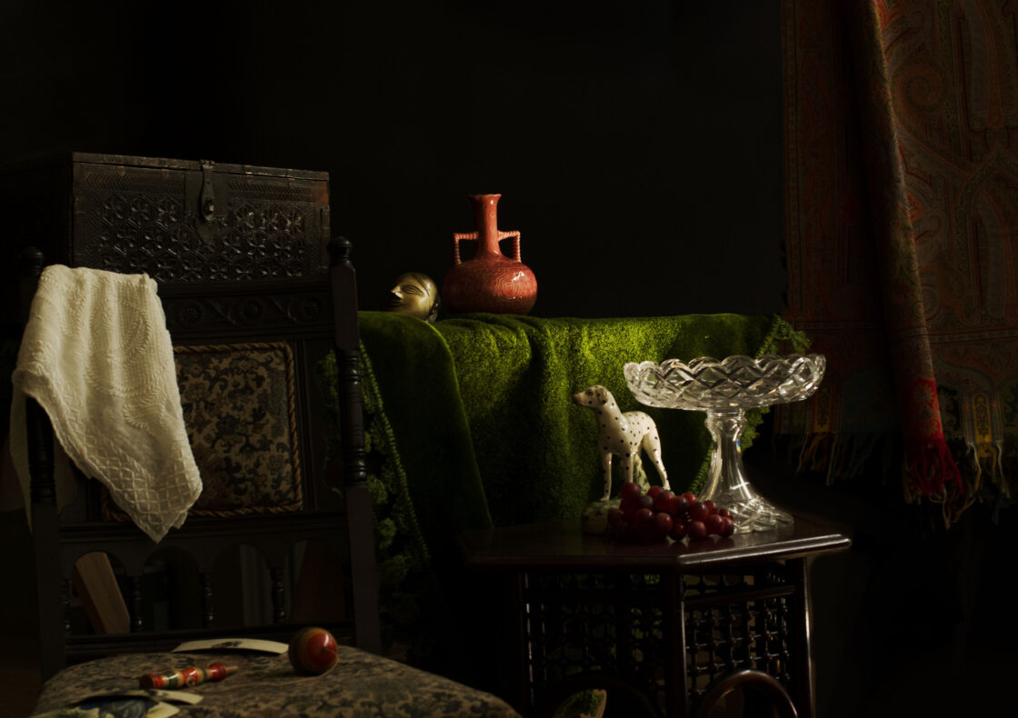A dimly lit still‐life setting evoking a 19th-century parlour: in the foreground, an ornate wooden chair upholstered in dark damask holds a folded white fabric over its back. Beside it, a small square table displays a crystal pedestal bowl, a spotted ceramic dog figurine, and a cluster of deep red grapes. In the back is another table draped in moss green fabric with a pink vase. To the right, a richly patterned hangs against the shadowed wall.
