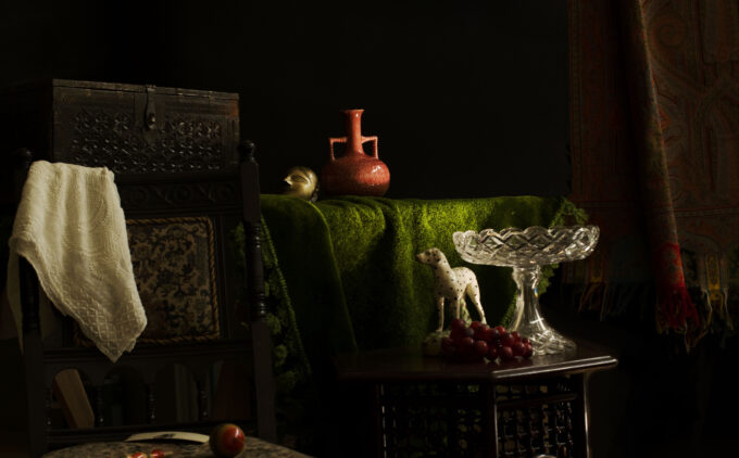 A dimly lit still‐life setting evoking a 19th-century parlour: in the foreground, an ornate wooden chair upholstered in dark damask holds a folded white fabric over its back. Beside it, a small square table displays a crystal pedestal bowl, a spotted ceramic dog figurine, and a cluster of deep red grapes. In the back is another table draped in moss green fabric with a pink vase. To the right, a richly patterned hangs against the shadowed wall.