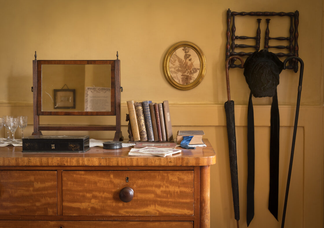 Detail of an interior featuring a wooden dresser with books, a mirror, sherry glasses, oval portrait on yellow walls and other period furnishings.