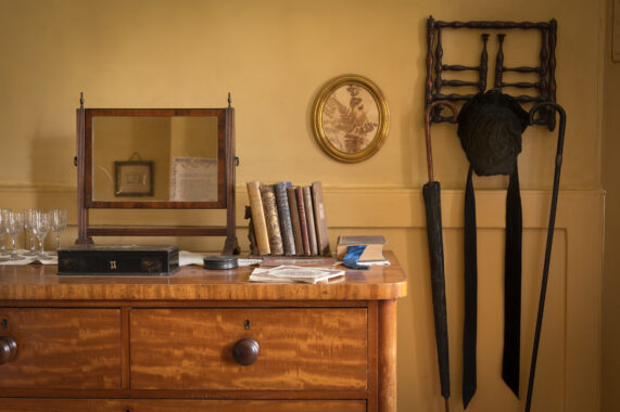 Detail of an interior featuring a wooden dresser with books, a mirror, sherry glasses, oval portrait on yellow walls and other period furnishings.