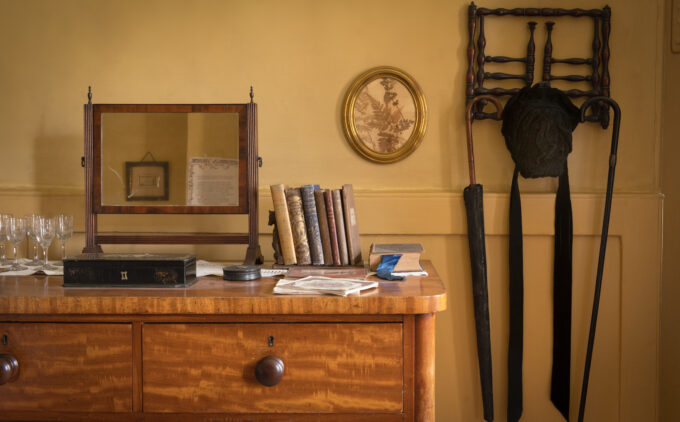 Detail of an interior featuring a wooden dresser with books, a mirror, sherry glasses, oval portrait on yellow walls and other period furnishings.
