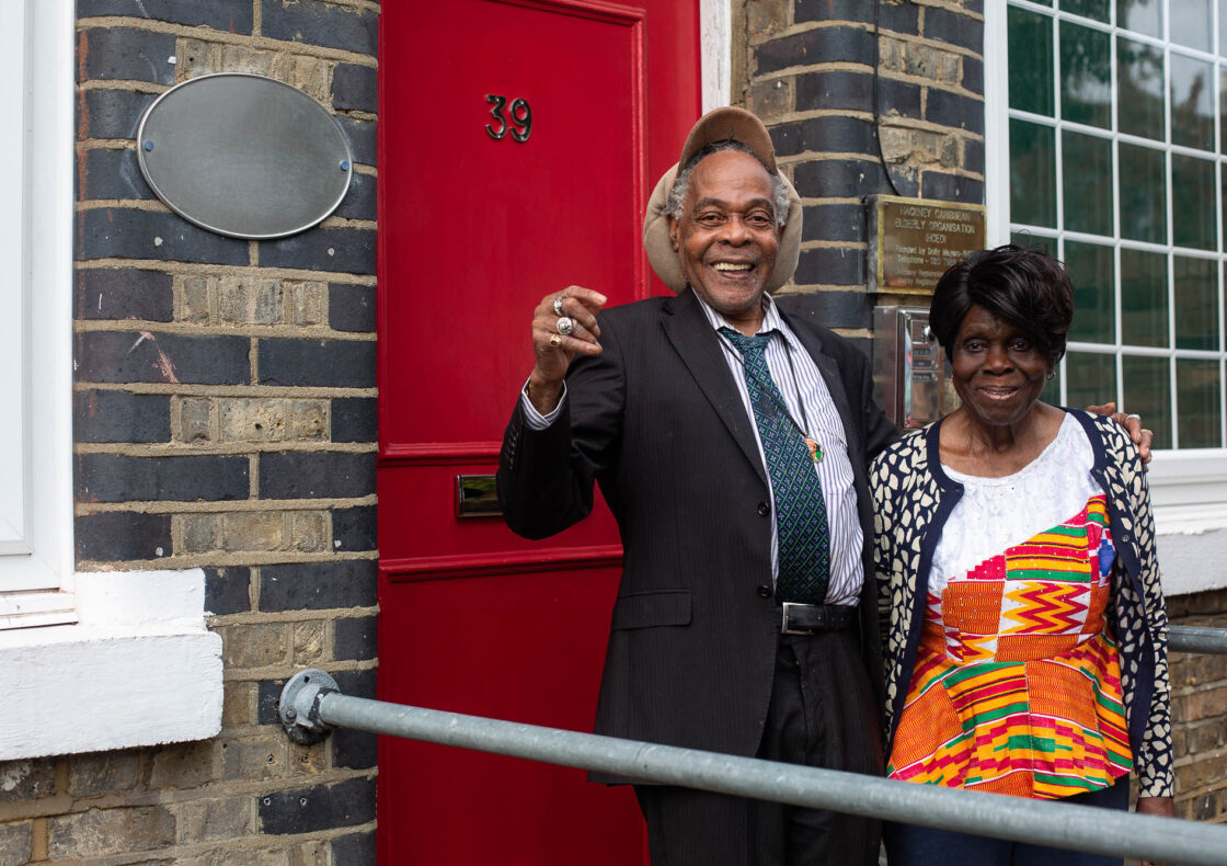 An elderly black gentleman and black woman smile at the camera in front of a red door and brick building. A sign reads "Hackney Caribbean Elderly Organisation"