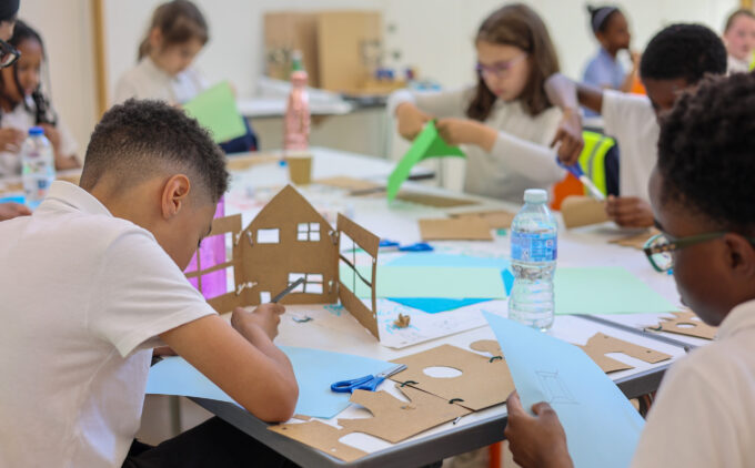 School children sit around a table working on crafts and cutting paper to make paper houses