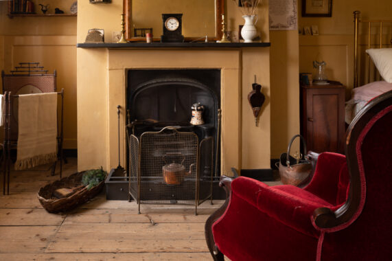 Room interior with a large fireplace, red velvet chair, wooden floors, furnishings including a mantel clock, brass candlesticks, and a four-poster bed.