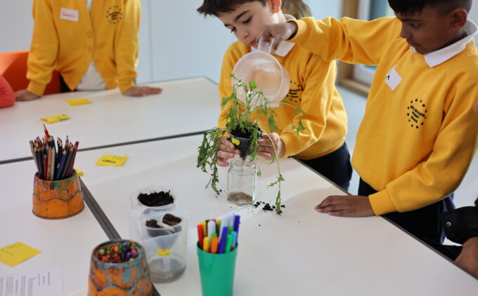 Two children in yellow uniforms water a plant at a white table inside, while pencils and containers are scattered around.