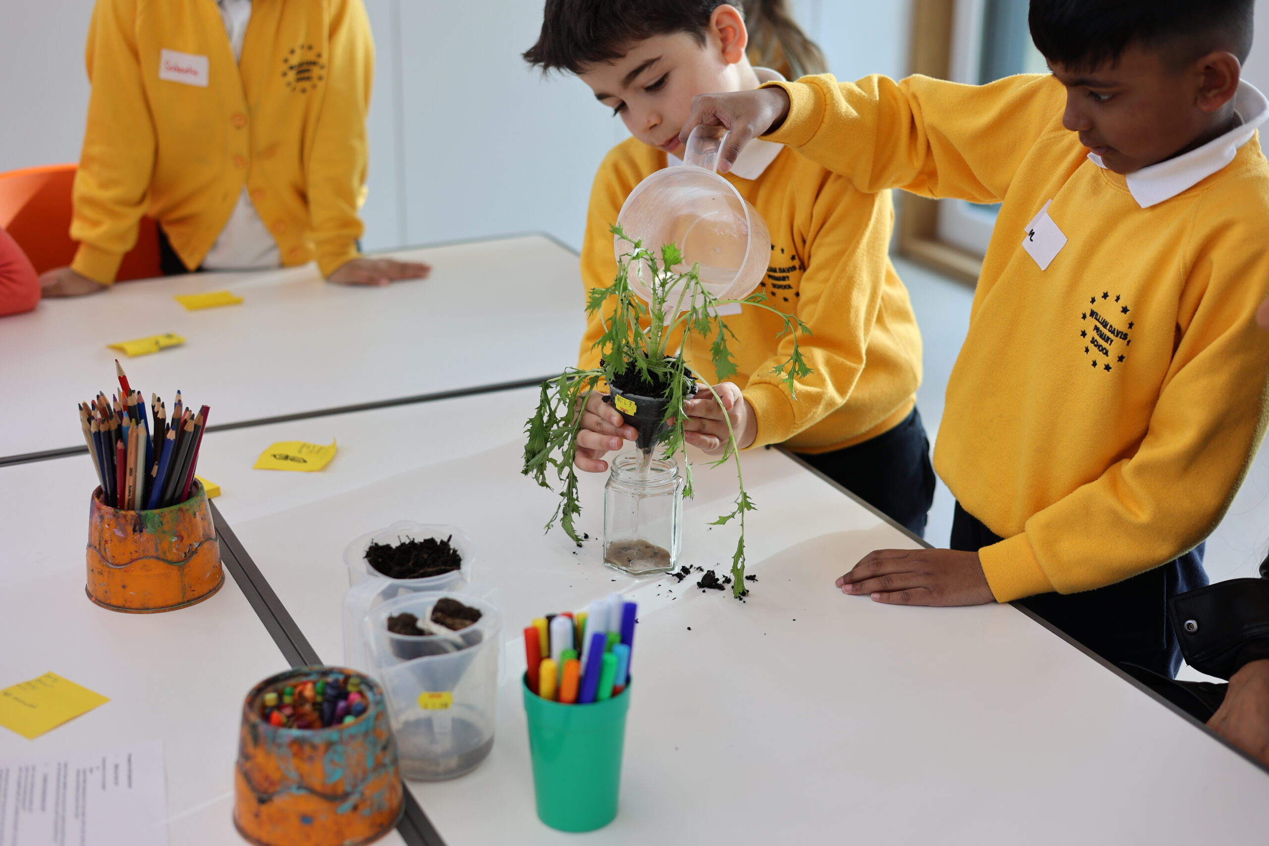 Two children in yellow uniforms water a plant at a white table inside, while pencils and containers are scattered around.