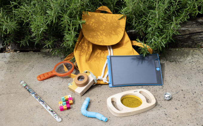 A yellow backpack is surrounded by fidgit toyds, a magnifying glass,a kaleidoscope camera, a glitter wand and a magic drawing board.
