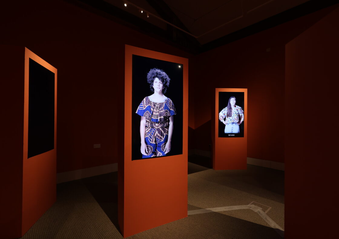 A dimly lit gallery room painted in deep terracotta, with several free-standing orange pillars arranged in a staggered layout. Each pillar holds a vertical digital screen displaying a life-sized portrait of a person against a black background. White tape on the floor marks visitor pathways through the installation.