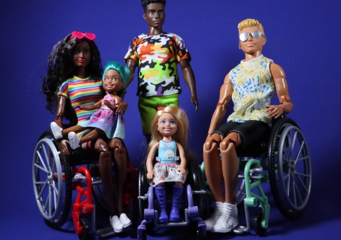 Group of five fashion dolls against a blue background. Two adult dolls sit in wheelchairs on both sides, one on the left holding a child doll on their lap. A young doll in a small wheelchair sits in the middle. The fifth doll is standing behind these four dolls.