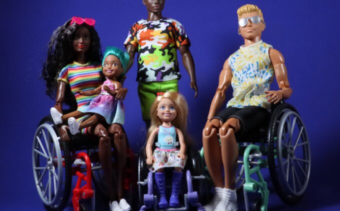 Group of five fashion dolls against a blue background. Two adult dolls sit in wheelchairs on both sides, one on the left holding a child doll on their lap. A young doll in a small wheelchair sits in the middle. The fifth doll is standing behind these four dolls.