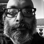 Black and white photo of a man with a beard and glasses staring at the camera.